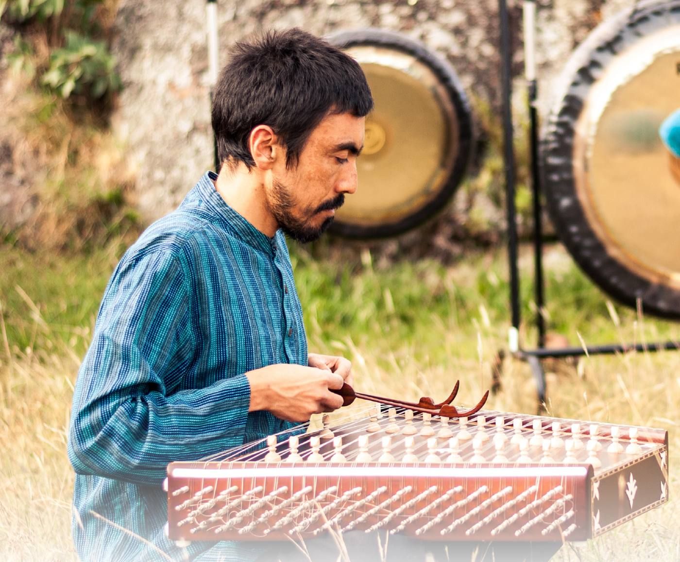 Músico tocando santur en el pasto, con dos gongs detrás